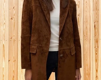 Op de afbeelding: Een bruine suède blazer met een ingekeepte revers, lange mouwen en twee klepzakken aan de voorzijde. Het jasje wordt gesloten met twee zichtbare knopen aan de voorzijde en wordt gedragen over een wit shirt met ronde hals en een donkere broek.