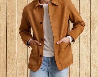 Op de afbeelding: Een bruin suède jasje met een knoopsluiting en twee voorzakken. Het jasje wordt gedragen over een wit shirt en een blauwe spijkerbroek. Het jasje heeft een klassieke kraag en lange mouwen. De achtergrond is een lichtbruine houten wand.