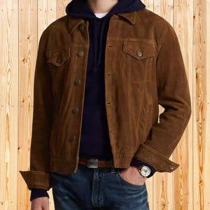 Puede incluir: Chaqueta de ante marrón con botones, dos bolsillos en el pecho y cuello. La chaqueta se lleva sobre una sudadera con capucha azul marino y un jersey azul oscuro. La persona lleva vaqueros azules y un reloj.