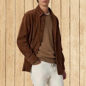 May include: A brown suede shirt jacket with button closures, worn over a tan sweater and paired with white pants. The jacket has a classic collar and long sleeves. The outfit is casual and stylish.