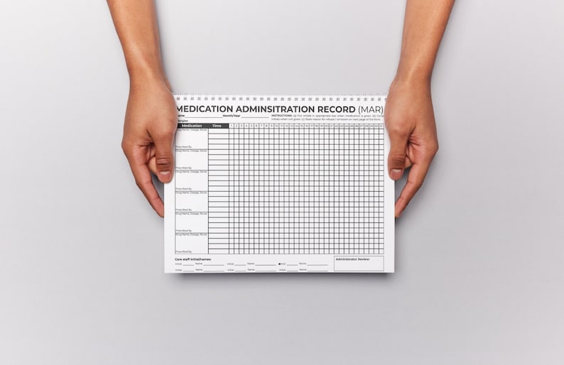 Puede incluir: Un bloc de notas de registro de administraci&oacute;n de medicamentos (MAR) es sostenido por dos manos. El bloc de notas es blanco con l&iacute;neas de cuadr&iacute;cula y texto en negro. El texto "MEDICATION ADMINISTRATION RECORD (MAR)" est&aacute; impreso en la parte superior.
