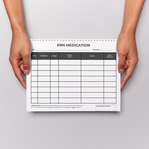Puede incluir: Un bloc de notas blanco con espiral con el texto "PRN MEDICATION" en la parte superior. El bloc de notas tiene un dise&ntilde;o de cuadr&iacute;cula para los detalles de la medicaci&oacute;n. Sostenido por dos manos sobre un fondo gris.