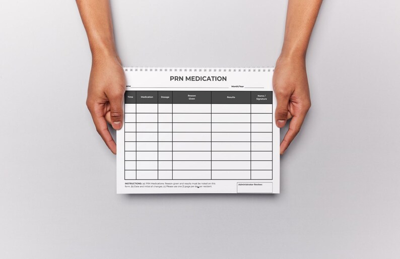 Puede incluir: Un bloc de notas blanco con espiral con el texto "PRN MEDICATION" en la parte superior. El bloc de notas tiene un dise&ntilde;o de cuadr&iacute;cula para los detalles de la medicaci&oacute;n. Sostenido por dos manos sobre un fondo gris.