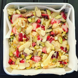 Puede incluir: Un molde para hornear blanco lleno de una mezcla de fresas picadas, pistachos, almendras y chocolate blanco. La mezcla está cubierta con una salsa blanca.