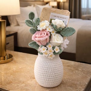 Puede incluir: Un jarrón blanco de ganchillo lleno de un ramo de flores hechas a mano. Las flores incluyen rosas rosas, rosas blancas y pequeñas flores blancas y amarillas, con hojas verdes. Un papel con el texto "PICNIC SEASON" está en el ramo.