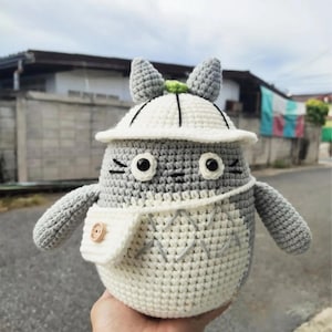 Puede incluir: Un peluche de Totoro hecho a mano con ganchillo en gris y blanco, que lleva un sombrero blanco con un borde negro y una pequeña bolsa blanca con un botón de madera. El peluche tiene ojos negros y boca cosida.