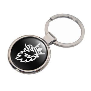 May include: A round, silver-toned keychain with a black insert featuring a white graphic design of a crowned creature. The keychain has a split ring for attaching keys. The design is centered on the black background.