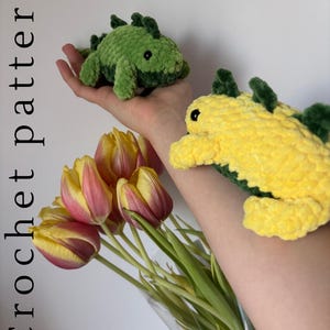 May include: Two crocheted dinosaur toys, one green and one yellow, with dark green spines. The toys are held near a bouquet of yellow and pink tulips. The text "Crochet pattern" is vertically aligned on the left side of the image.