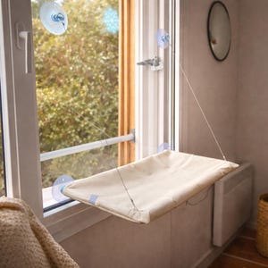 Peut inclure: Un perchoir de fenêtre pour chat beige, suspendu par des ventouses. Le lit rectangulaire en tissu doux est suspendu devant une fenêtre, offrant une vue sur l'extérieur. Le perchoir est conçu pour offrir un endroit confortable pour que le chat se détende.