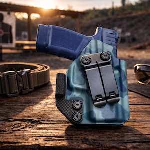 May include: A blue handgun in a camouflage holster rests on a wooden surface, alongside a brown belt with a metal buckle and black sunglasses. The holster has a black clip and carbon fiber accents. The background includes a shooting range.