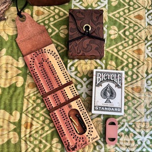 May include: A wooden cribbage board with leather accents and a skunk graphic labeled "I Smell A Skunk," a brown embossed leather card case with a brass closure, a deck of Bicycle standard playing cards, and a small rectangular leather piece.