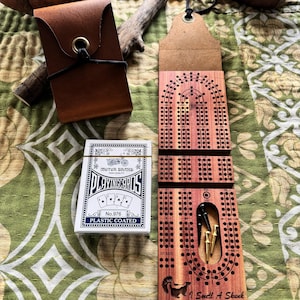 May include: A wooden cribbage board with a skunk illustration and the text "I Smell A Skunk" next to a deck of Motor Brand playing cards. A brown leather card holder and a leather pouch are also visible.