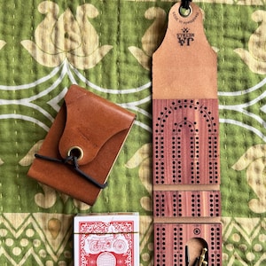 以下が含まれることがあります： 茶色の革製カードケースと、赤と白のデザインのトランプが、木製のクリベッジボードの隣にあります。クリベッジボードは革のトップで、「I Smell A Skunk」の文字が入っています。