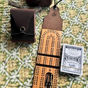 May include: A wooden cribbage board with a skunk graphic and the text "I Smell A Skunk". The board is open, revealing metal pegs. A deck of playing cards and a leather card holder are also present.