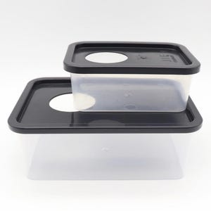 Puede incluir: Dos recipientes transparentes de plástico para guardar alimentos con tapas negras. Las tapas tienen una abertura circular. Los recipientes son rectangulares y apilables. Sobre fondo blanco.