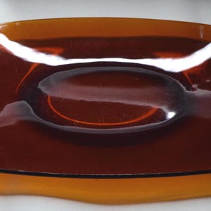 May include: A rectangular amber-colored glass tray with rounded corners and a smooth, polished surface. The tray features a central oval indentation and a translucent, deep orange hue that catches the light, creating bright reflections across its top.