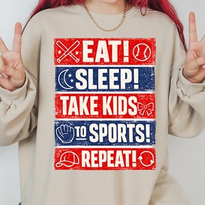 May include: Beige sweatshirt with a red, white, and blue graphic. The text reads "EAT! SLEEP! TAKE KIDS TO SPORTS! REPEAT!" with baseball and sports-themed icons. A casual, comfortable top for sports fans.