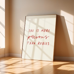 Puede incluir: Una impresión artística enmarcada con el texto "SHE IS MORE precious THAN RUBIES" en escritura roja sobre un fondo blanquecino. El marco es dorado y la impresión está apoyada contra una pared.