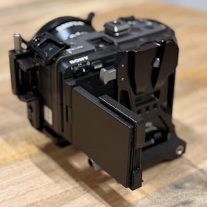 May include: A black Sony digital camera body equipped with a protective metal camera cage. The device features a side-mounted articulating screen that is folded outward, multiple mounting points, and a top-mounted hot shoe for external accessories.