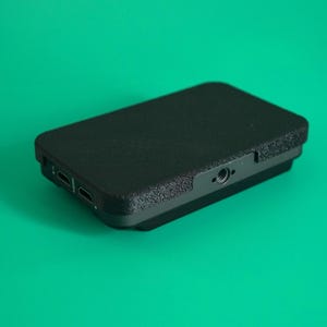 May include: A black rectangular device with a textured surface, possibly an external hard drive or media player, sits against a solid green background. The device has ports on one side and a small hole on the other.