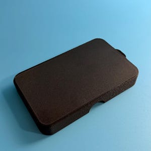 May include: A rectangular, black case with a textured surface. The case has rounded corners and a small notch on one side. The object is set against a solid blue background.