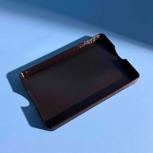 May include: A black rectangular phone case with a textured surface. The case has a raised edge and a cutout for the camera. The case is sitting on a blue surface.
