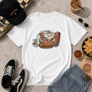 May include: White t-shirt featuring a colorful cartoon illustration of a kebab vendor. The graphic includes the word "KEBAB!" and the phrases "¿PARA LLEVAR?!" and "PARA TOMAR AQUI". The image also shows a black baseball cap, sunglasses, and a pair of black and white sneakers.