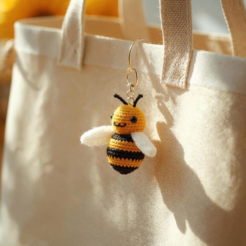 May include: A crocheted bee keychain with a yellow head and body, black stripes, and white wings. The bee is attached to a gold-colored ring, hanging from a beige tote bag. The bee has black antennae and a smiling face.