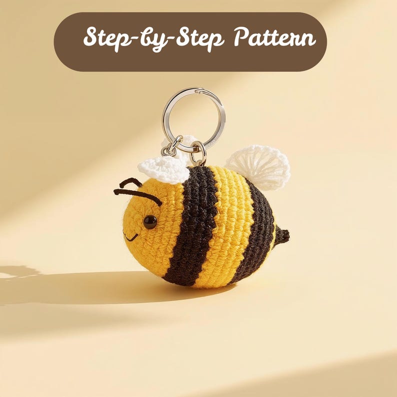 May include: A crocheted bee keychain with yellow and black stripes, white wings, and a small white hat. The keychain has a silver ring. The text "Step-by-Step Pattern" is at the top.