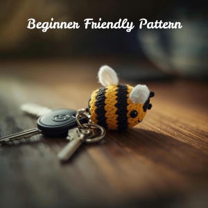 May include: A handmade crochet bee keychain with yellow and black stripes, white wings, and a smiling face. The keychain is attached to a set of keys. The text "Beginner Friendly Pattern" is displayed above the bee.