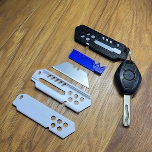 May include: A selection of utility knife parts and a car key are displayed on a wooden surface. The components include a black and a light grey knife handle, a blue blade holder, and several silver utility knife blades. The car key is black.