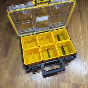 May include: A clear and yellow plastic tool organiser with the text "TOUGH SYSTEM" on the lid. The organiser has a black base with six yellow storage compartments and a black handle.