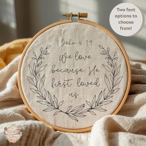 May include: Embroidered hoop art with the text "1 John 4:19 We love because He first loved us." The text is in a black script font, surrounded by a black floral wreath. A small, round text bubble says "Two font options to choose from!"