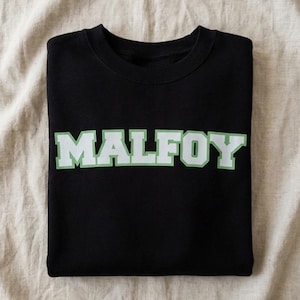 Könnte beinhalten: Schwarzes Sweatshirt mit dem Wort "MALFOY" in weißen Blockbuchstaben mit grünem Rand. Das Sweatshirt ist auf einem beigefarbenen Stoff gefaltet.
