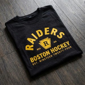 Könnte beinhalten: Schwarzes T-Shirt mit den gelben Worten "RAIDERS BOSTON HOCKEY". Das Shirt enthält den Text "EST. NINETEEN TWENTY-FOUR" und ein stilisiertes "R"-Logo.