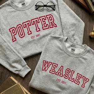 Può includere: Due felpe grigie con scritte rosse. Una dice "POTTER EST. 1997" e l'altra "WEASLEY EST. 1997". Un paio di occhiali rotondi neri sono sulla prima felpa. Libri sullo sfondo.