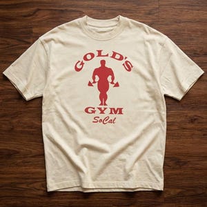 Könnte beinhalten: Ein cremefarbenes Kurzarm-T-Shirt mit einem roten Grafikdruck auf der Brust. Das Design zeigt den Schriftzug "GOLD'S GYM SoCal" um die Silhouette einer muskulösen Figur, die Hanteln hält, zentriert auf der Vorderseite des Kleidungsstücks.