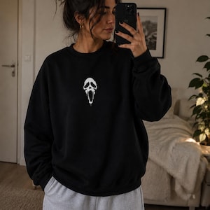 Könnte beinhalten: Ein schwarzes Langarm-Sweatshirt mit Rundhalsausschnitt und einer kleinen, mittig platzierten weißen Grafik eines stilisierten, schmelzenden Geistermasken-Motivs. Das Sweatshirt wird mit einer hellgrauen Jogginghose zu einem legeren Set kombiniert.