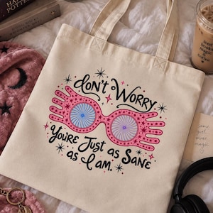 Op de afbeelding: Een beige canvas draagtas met een grafische print van roze brilmonturen met blauwe spiraalvormige glazen. De tekst luidt "Don't worry, you're just as sane as I am", omringd door kleine roze en zwarte sterren en decoratieve krullen.