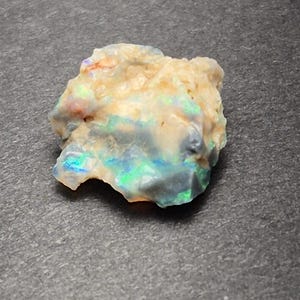 May include: A raw opal gemstone with a creamy white base. The opal exhibits flashes of blue, green, and orange iridescence. The stone has an irregular shape, highlighting its natural, unpolished form. The opal is a beautiful, natural stone.