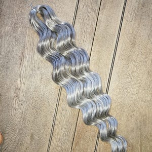 May include: A bundle of wavy, silver-grey hair extensions with a subtle blue hue. The hair is styled in loose waves and is secured at the top with a metal clip. The extensions are lying on a wooden surface.