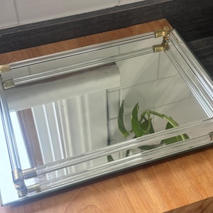 May include: A rectangular mirrored tray with clear acrylic sides and brass corner accents. The tray sits on a wooden base. The mirror reflects a plant and a white tiled wall.