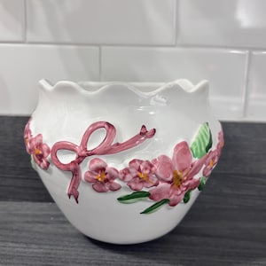 Peut inclure: Pot de fleurs en céramique blanche avec un bord festonné, orné de fleurs roses, de feuilles vertes et d'un nœud rose. Le motif floral est en relief, créant un effet tridimensionnel. Le pot est posé sur un fond neutre.