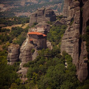 May include: A picturesque view of a monastery atop a large rock formation. The building has a red tiled roof and is surrounded by green trees. The landscape features rugged cliffs and a distant mountain range.