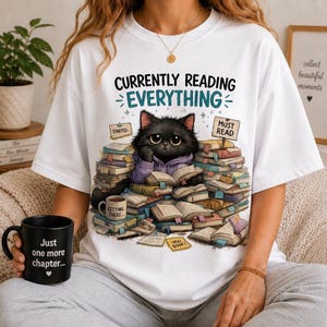 May include: A white t-shirt featuring a graphic of a black cat wearing a purple hoodie, sitting amidst a large pile of open books. The text reads "CURRENTLY READING EVERYTHING," "STARTED," "MUST READ," "GETTING THERE," and "YES BOOK."