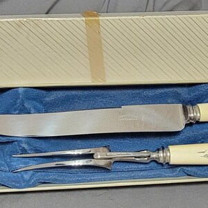 May include: A vintage carving set in a box. The set includes a large knife and a carving fork with cream-coloured handles featuring a black landscape design. The set rests on a blue velvet-like fabric inside a cream-coloured box.