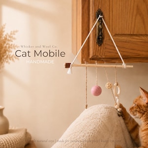 May include: A handmade cat mobile with a wooden dowel and hanging toys. The mobile includes a pink felt ball, a woven ball, and a wooden ring. The text "Cat Mobile" and "HANDMADE" are visible.