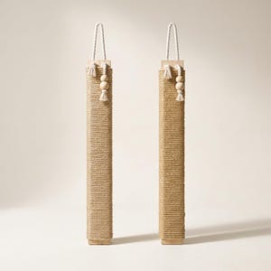 May include: Two vertical rectangular scratching posts wrapped in tan sisal rope. Each post features a light-colored wooden top and bottom cap, a white hanging cord, and a decorative accent consisting of two wooden beads and a small white tassel.