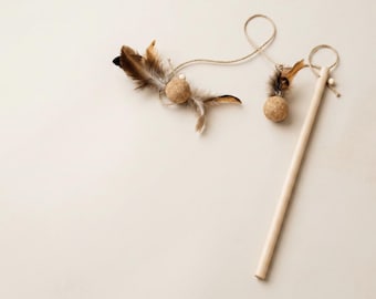 May include: A cat toy consisting of a smooth wooden rod attached to a long piece of twine. The twine features two small, textured cork balls adorned with brown and tan feathers, along with small wooden beads secured at the ends of the string.