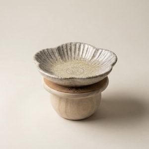 Puede incluir: Un plato de cerámica con forma de flor y esmalte moteado en beige y gris, con un sello circular central. El plato descansa sobre un posavasos de corcho redondo encima de una base de madera clara y lisa con el texto W & W CO impreso en el corcho.